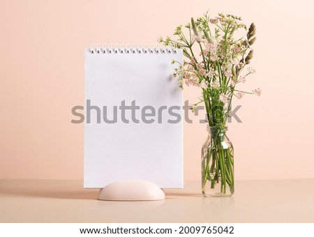 The planner's blank clean notepad stands on the table in vase with delicate green flowers. Mock up for text
