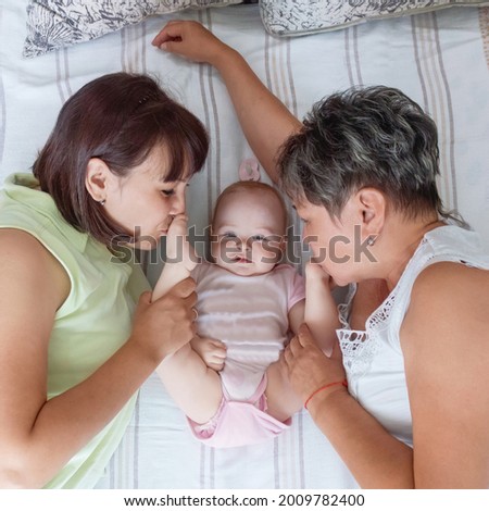Grandmother and mother kisses little baby girl feets smiling cheerfully  at kid on the whit bed at home.
