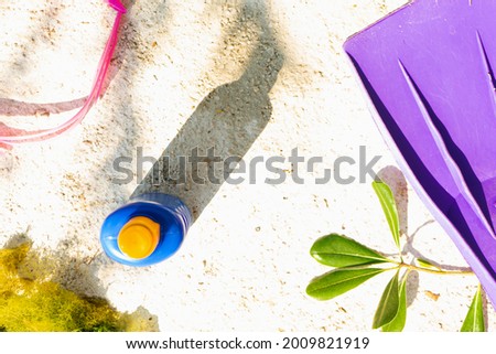 A flat lay of sunscreen cream blue bottle, swimming goggles, sea weed, pier branch leaves with purple flippers under summer shade. Minimal holiday vacation arrangement.