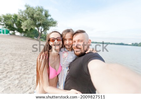 
Family of mom dad and daughter hug and take selfie on the beach. Morning, summer. River.