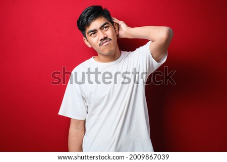 Young handsome man with mustache wearing white t-shirt standing over red background confuse and wonder about question. Uncertain with doubt, thinking with hand on head. Pensive concept.
