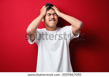 Young handsome asian man wearing white t-shirt standing over isolated red background with hand on head for pain in head because stress. Suffering migraine.