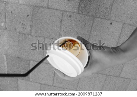A two euro coin at the bottom of a paper coffee cup. Leftover coffee and a plastic tube. Hold a glass in your hand against a background of paving stones.