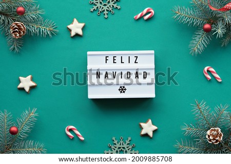 Light box with text Feliz Navidad - Merry Christmas in Spanish language. Xmas background, top view on fir twigs decorated with red glass toys, decorations on vibrant turquoise textile, tablecloth.