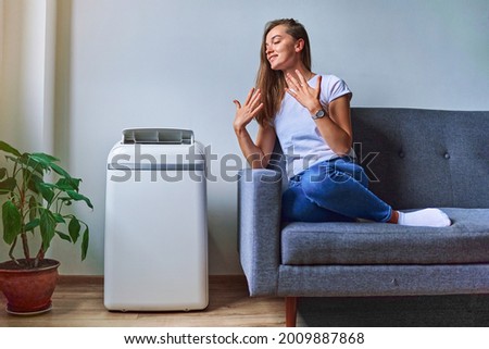Young casual woman sitting on a sofa and using floor air conditioner for cooling in hot summer weather. Fresh and clean air at home Royalty-Free Stock Photo #2009887868