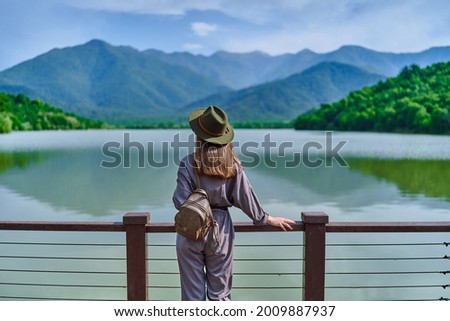 Traveler girl standing alone on edge of pier and staring at lake and mountains. Enjoying happy freedom moment life and serene quiet peaceful atmosphere in nature. Back view
