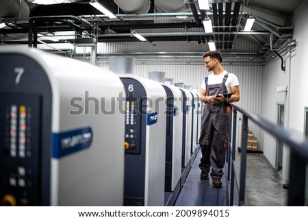 Printing house interior with modern offset print machine and operator in working uniform checking quality and controlling process of print. Royalty-Free Stock Photo #2009894015