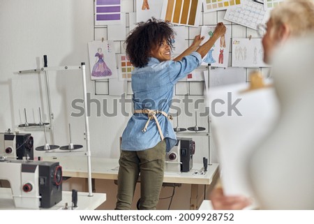 African-American fashion designer puts dresses models on wall looking at mature colleague