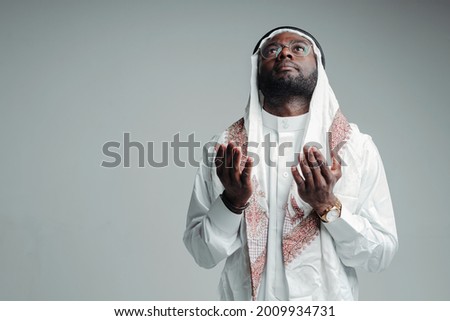 Arab muslim man praying in mosque stretching palms up and looking up while standing on gray background Royalty-Free Stock Photo #2009934731