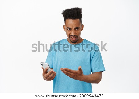 Annoyed african american male model, holding smartphone, furrow eyebrows and pointing at mobile phone with upset grimace, complaining, bothered by stupid message, white background