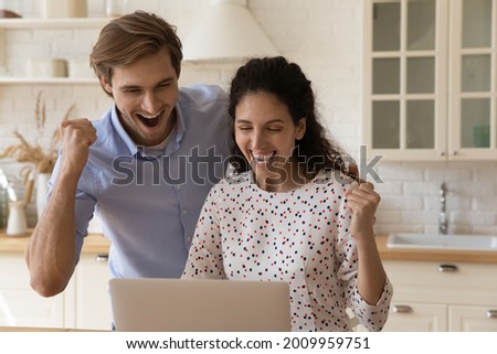 Overjoyed young family couple stand at kitchen look at laptop screen celebrate receiving loan approval from bank. Excited spouses real estate agency clients get email offering house of dream for sale Royalty-Free Stock Photo #2009959751