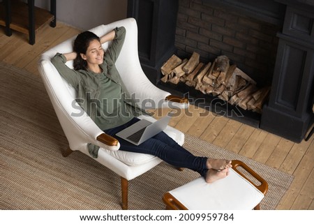 Peaceful latina woman lean back in comfy chair with hands over head take break in work with laptop relax at modern living room near fireplace. Calm young lady enjoy rest at home breath clean fresh air Royalty-Free Stock Photo #2009959784