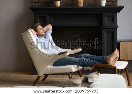 Carefree young male homeowner relaxing barefoot in capacious armchair at luxury apartment by modern fireplace. Serene single millennial male rest with hands behind head at rich comfortable living room Royalty-Free Stock Photo #2009959793