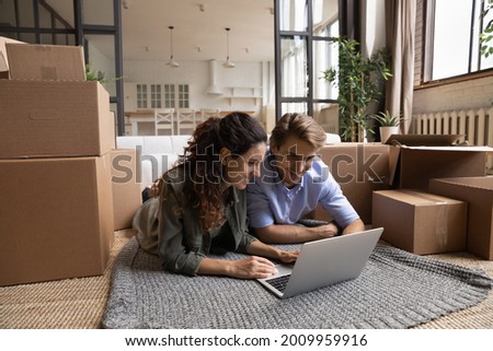 Enthusiastic millennial spouses buyers renters of new flat lie on carpet on floor among boxes packages at moving day use laptop. Inspired couple in love choose domestic interior design on pc online Royalty-Free Stock Photo #2009959916