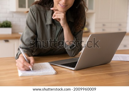 Elearning. Close up of smiling young female remote student watch training lecture on laptop pc take notes at paper workbook by hand. Millennial lady get distant education write thesis of online lesson