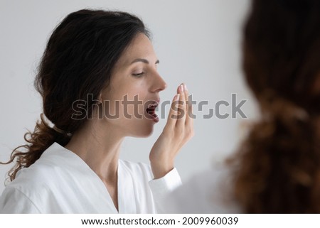 Fresh like wind blow. Millennial hispanic woman wearing bathrobe standing by mirror holding palm by opened mouth checking breath. Young latin female testing breathing smelling air holding palm by lips Royalty-Free Stock Photo #2009960039