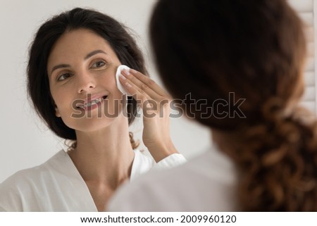 Caring for skin. Pretty millennial hispanic woman do everyday facial skincare procedure cleansing face with cosmetic lotion using cotton sponge. Young female remove makeup at evening by mirror at home Royalty-Free Stock Photo #2009960120