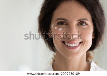 Close up portrait of smiling young latin female face with cosmetic cream moisturizer drops on skin under eyes. Happy millennial lady look at camera introduce new antiage creme serum lotion. Copy space Royalty-Free Stock Photo #2009960189