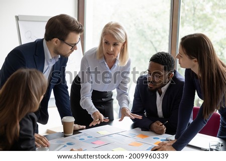 Mature business teacher training group of employees, teaching to work with paper reports, analyze marketing charts, using sticky notes. Mentor explaining scrum method tools to employees Royalty-Free Stock Photo #2009960339
