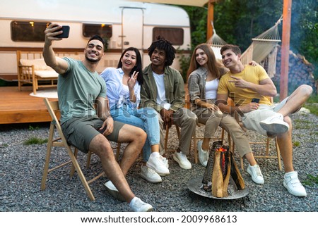 Cheerful diverse young friends taking selfie near campfire in evening, using smartphone, sitting next to RV during camping trip, spending summer vacation outside, having fun together