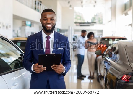 Portrait Of Handsome Black Car Salesman In Suit Posing At Workplace In Auto Showroom, Young Dealership Center Manager With Clipboard In Hands Helping Spouses To Purchase New Automobile, Free Space Royalty-Free Stock Photo #2009968772