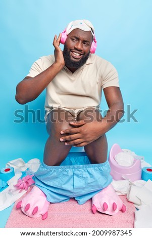 Vertical shot of happy dark skinned man enjoys listening music uses wireless headphones dressed in domestic clothes feels comfortable while sitting on toilet bowl isolated on blue background