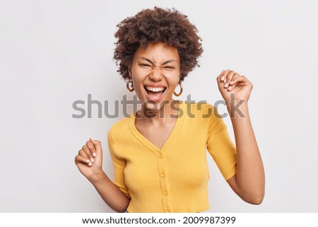 Beautiful happy young woman with curly hiar feels free and pleased raises hands up has fun dances joyful against white background expresses positive authentic emotions wears casual yellow jumper