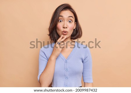 Confused shocked brunette Asian woman reacts on something with wondered frightened expression wears blue jumper looks startled stares at promo logo wears blue jumper isolated over beige wall Royalty-Free Stock Photo #2009987432