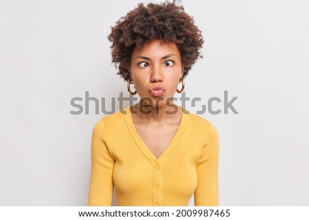 Photo of funny curly haired pretty Afro American woman keeps lips folded  makes grimace at camera foolishes around wears yellow jumper isolated over white background has fun alone