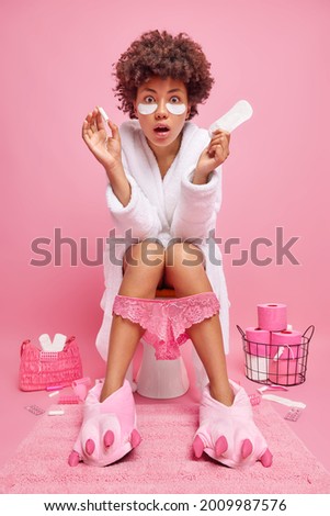 Photo of shocked curly haired young woman holds sanitary napkin and tampon chooses hygiene product for menstruation period undergoes skin care procedures at home poses on toilet in lavatory.