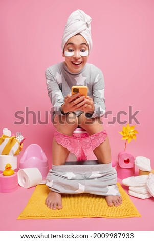 Pretty young Asian woman dressed in nightwear applies beauty patches uses mobile phone while sitting on toilet wears towel wrapped over head isolated over pink background. Domestic activities