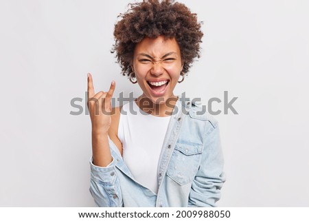 Happy crazy curly haired woman shows rock and roll gesture heavy metal sign enjoys favorite music on party has fun squints face exclaims from joy wears denim shirt isolated over white background.