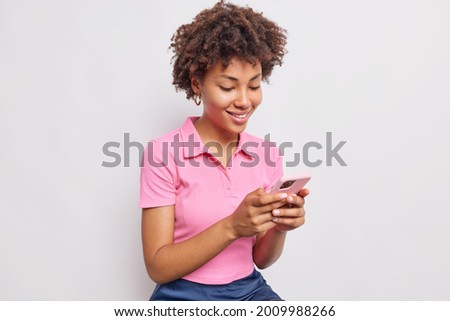 Smiling good looking woman with curly hair uses mobile phone for sending text messages and scrolling newsfeed dressed in casual pink t shirt isolated over white background. Technology concept