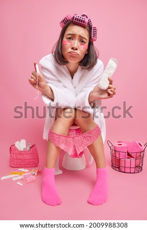 Unhappy Asian woman has menses suffers from period cramps sits on toilet bowl holds tampon and sanitary napkin looks sadly makes hairstyle undergoes beauty treatments dressed in domestic clothes