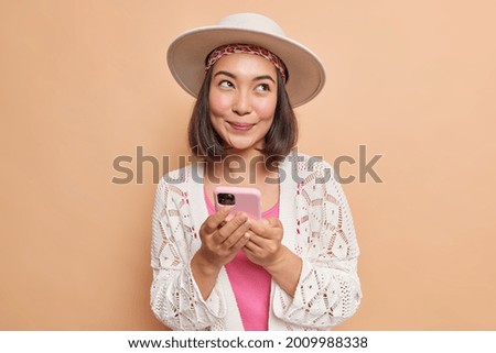 Pleased dreamy young Asian woman holds modern smartphone in hands uses cellular application for chatting online looks away wears stylish hat white knitted shawl isolated over brown background