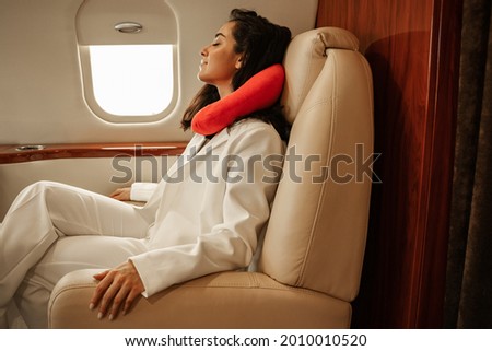 Rest on the plane. A young girl in a white business suit is napping while sitting by the porthole in a leather chair in a business jet using a red neck pillow for travel Royalty-Free Stock Photo #2010010520