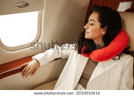 Rest in the plane. A young girl in a white suit is resting and looking in the window of a business plane, using a red neck pillow for travel Royalty-Free Stock Photo #2010010616