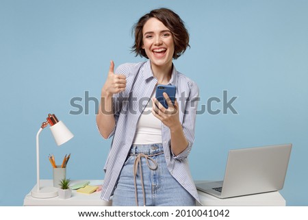 Young happy fun secretary employee business woman in casual shirt work stand at white office desk with pc laptop hold mobile cell phone show thumb up gesture isolated on pastel blue background studio. Royalty-Free Stock Photo #2010041042