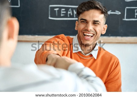 Young happy laughing Indian latin Hispanic creative startup coworker student giving fist bump to classmate partner committing successful project result in office classroom at desk near blackboard. Royalty-Free Stock Photo #2010062084