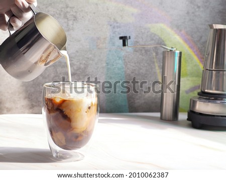 Hand in sterilized gloves pouring milk over ice balls with espresso in double wall insulated glass on white background and rustic grey wall with rainbow light reflection. Refreshing summer drink theme