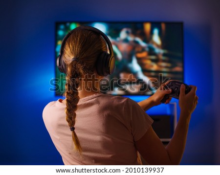 A young blonde woman is enthusiastically playing a video game. She has headphones on her head and a joystick in her hands. Soft neon light from the monitor floods the room.