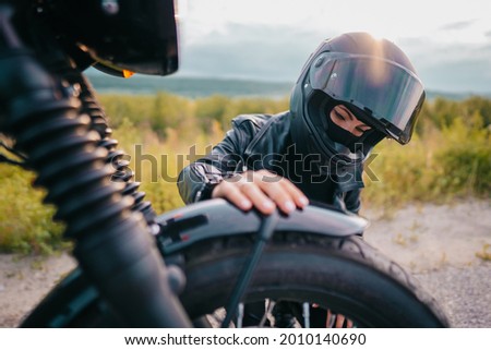 Portrait of confident motorcyclist woman in helmet sitting near bike. Young driver biker outdoors at sunset.