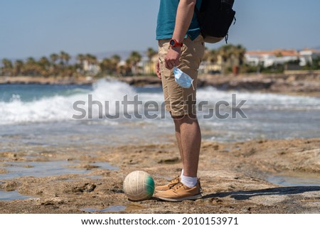 Theme of lockdown, quarantine of spoiled vacation due to travel restrictions cornavirus pandemic. Unrecognizable man stands with mask in hand and volleyball against background of sea on rocky shore. Royalty-Free Stock Photo #2010153971