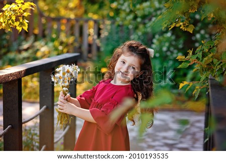 A girl in a red dress on a wooden bridge in a summer garden. Portrait of a little girl with a bouquet of daisies.