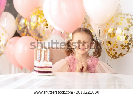 Make a wish.Child girl in festive dress closed eyes with strong emotions making birthday wish before blowing out candles on birthday cake. Balloons background.Child's birthday
