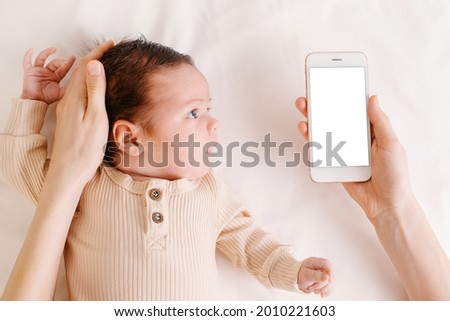 Baby newborn on white bed, woman holding mobile phone with white screen in hand top view.Technology and childhood concept