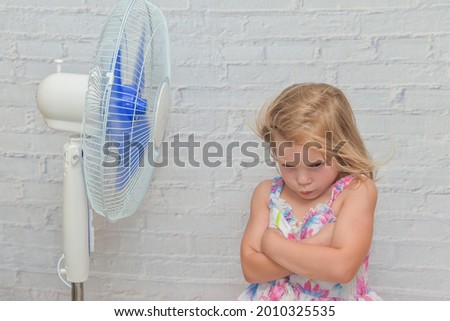 the girl child in front of an electric fan enjoys the flow of cool wind on a hot summer day Royalty-Free Stock Photo #2010325535