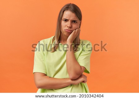 Sad bored young woman in yellow tshirt keeps hands folded and feels tired isolated over orange background Looking at camera Royalty-Free Stock Photo #2010342404