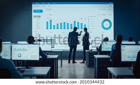 Two Traders Having a Meeting in a Modern Monitoring Office with Analytics Feed on a Big Digital Screen. Monitoring Room with Brokers and Finance Specialists Sit in Front of Computers. Colleagues Talk.