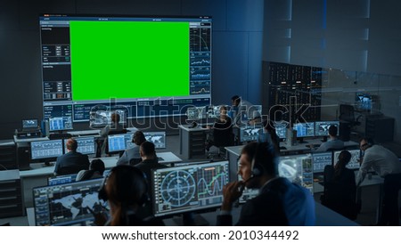 Big Green Screen Horizontal Mock Up in a Mission Control Center Room with Flight Director and Other Controllers Working on Computers. Team of Engineers Work in Monitoring Room Full of Displays. Royalty-Free Stock Photo #2010344492